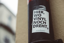 A black pole with a white poster attached to it. The poster contains bold black text in German that reads 'HIER, WO VINYL NOCH DREHT'. The background shows a blurred building facade with soft, natural lighting.