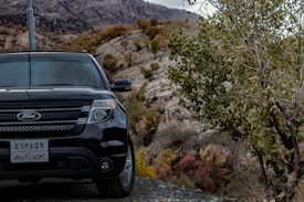 A black Ford SUV is parked on a rugged, mountainous terrain. The vehicle occupies the left side of the frame, while to the right, the sloping landscape, characterized by rocky hills and sparse trees, extends into the background. There is a pole with a wire cutting across the scene. The car's license plate has numbers and script in Arabic.