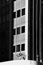 A black-and-white image depicts a high-rise building with a grid-like pattern of windows. A person wearing a harness and work gear is standing on a ledge, seemingly involved in maintenance or cleaning activity on the facade.