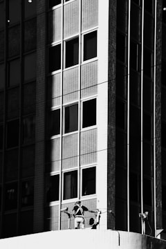 A black-and-white image depicts a high-rise building with a grid-like pattern of windows. A person wearing a harness and work gear is standing on a ledge, seemingly involved in maintenance or cleaning activity on the facade.
