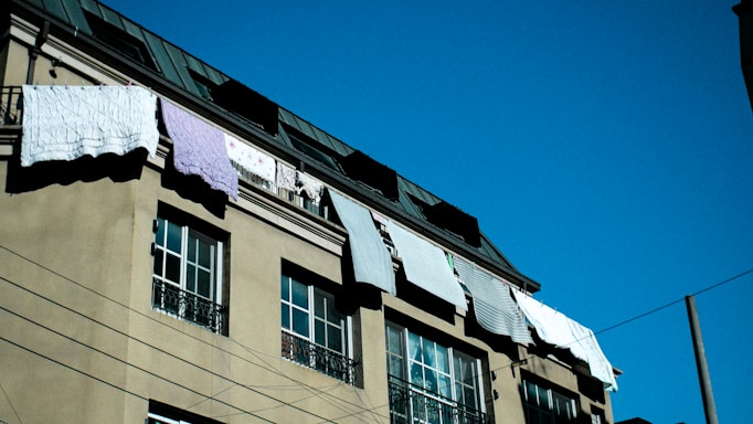 A residential building with several windows and a slanted roof has laundry hanging out to dry on a line. The sky is clear and deep blue, creating a contrast with the beige facade of the building.