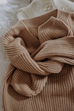 A beige knitted sweater with a textured pattern is casually laid out. The sweater is placed on top of a white knitted garment, possibly another sweater or blanket, both resting on a quilted white surface.