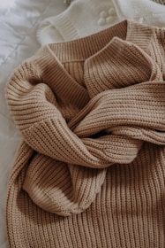 A beige knitted sweater with a textured pattern is casually laid out. The sweater is placed on top of a white knitted garment, possibly another sweater or blanket, both resting on a quilted white surface.