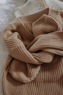 A beige knitted sweater with a textured pattern is casually laid out. The sweater is placed on top of a white knitted garment, possibly another sweater or blanket, both resting on a quilted white surface.