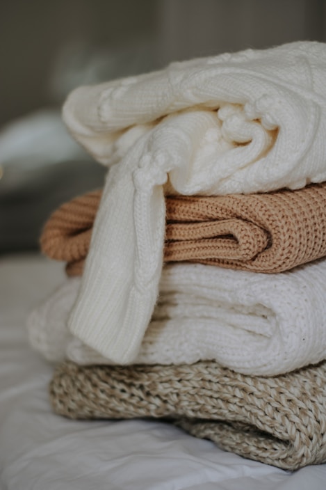 A neatly stacked pile of cozy knitted sweaters featuring various textures and patterns. The top sweater is a creamy white with thick, soft knitting, followed by a warm caramel-colored sweater. Below, there are additional cream and beige sweaters, all layered on a bed, suggesting comfort and warmth.