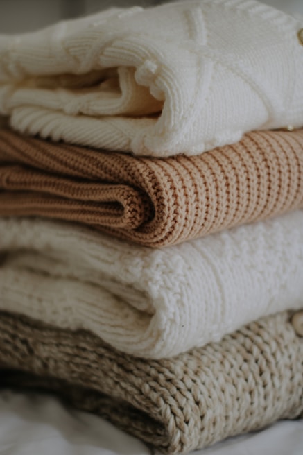 A stack of knitted sweaters in various neutral tones. The top sweater is a soft white with a textured pattern, followed by a light tan sweater, then another white one, and a knitted brown one at the bottom. The image conveys a cozy and warm atmosphere.