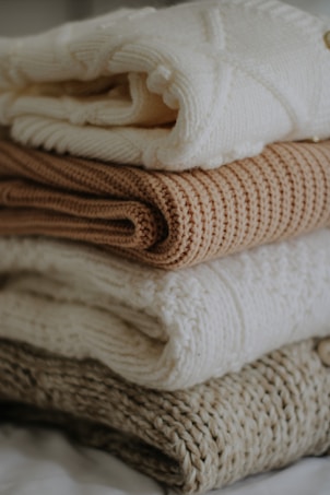 A stack of knitted sweaters in various neutral tones. The top sweater is a soft white with a textured pattern, followed by a light tan sweater, then another white one, and a knitted brown one at the bottom. The image conveys a cozy and warm atmosphere.