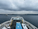 A ferry is traveling across a body of water with a cloudy sky overhead. Seagulls fly above the ferry and another ferry is visible in the distance. The deck is lined with a few parked vehicles, and the view shows a distant shoreline with buildings stretching across the horizon.