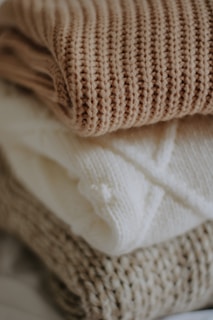 Three folded knitted sweaters stacked on top of each other in neutral tones. The top sweater has a brown ribbed pattern, the middle one is cream with a cable knit design, and the bottom sweater is beige with a thick knit texture.