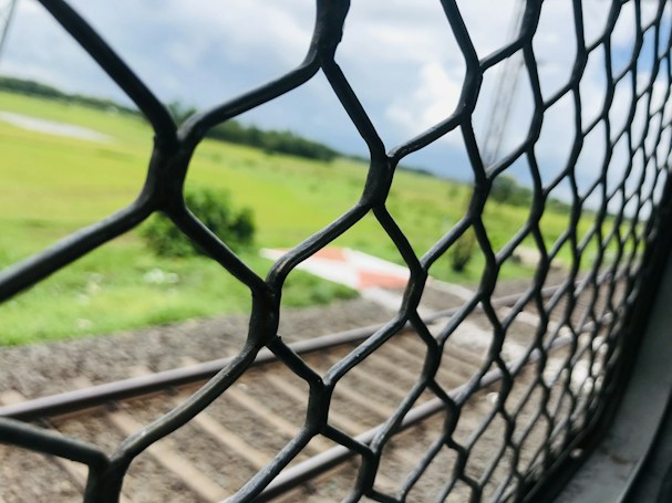 A chain-link fence in the foreground with blurred railway tracks and a vast green landscape in the background. The sky is partially cloudy, adding a contrast to the vibrant green fields.