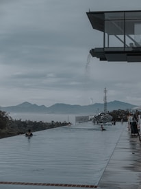 An infinity pool with a modern architectural design overlooks a scenic mountain landscape. The pool appears to blend seamlessly with the horizon, creating an illusion of endless water extending to the distant mountains. Several people are swimming, and a sculptural water feature adds a dynamic element to the scene. The surroundings have a serene and tranquil atmosphere enhanced by soft, muted lighting.