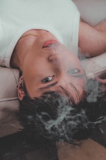 A person lies on their back facing upwards, partially covered by smoke. They have short hair and are wearing a white top with a gold necklace. The focus is on their face, which appears relaxed and contemplative.