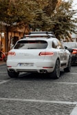 A white luxury SUV is parked on a cobblestone street, surrounded by trees and other vehicles. A bicycle can be seen to the left, and another car is partially visible near the rear of the SUV. The area has a calm, urban feel with subdued lighting.