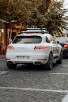 A white luxury SUV is parked on a cobblestone street, surrounded by trees and other vehicles. A bicycle can be seen to the left, and another car is partially visible near the rear of the SUV. The area has a calm, urban feel with subdued lighting.