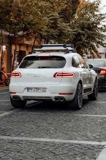 A white luxury SUV is parked on a cobblestone street, surrounded by trees and other vehicles. A bicycle can be seen to the left, and another car is partially visible near the rear of the SUV. The area has a calm, urban feel with subdued lighting.