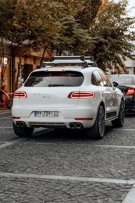A white luxury SUV is parked on a cobblestone street, surrounded by trees and other vehicles. A bicycle can be seen to the left, and another car is partially visible near the rear of the SUV. The area has a calm, urban feel with subdued lighting.
