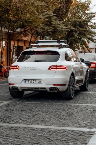 A white luxury SUV is parked on a cobblestone street, surrounded by trees and other vehicles. A bicycle can be seen to the left, and another car is partially visible near the rear of the SUV. The area has a calm, urban feel with subdued lighting.