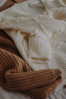 A collection of knitted sweaters in beige and cream colors, overlapping each other. The textures are prominently ribbed and feature a cozy pattern.