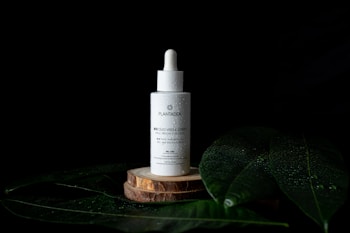 A dropper bottle labeled 'PLANTAEA' is placed on a wooden platform, surrounded by large green leaves with water droplets. The setting is against a dark background, emphasizing the product in the center.