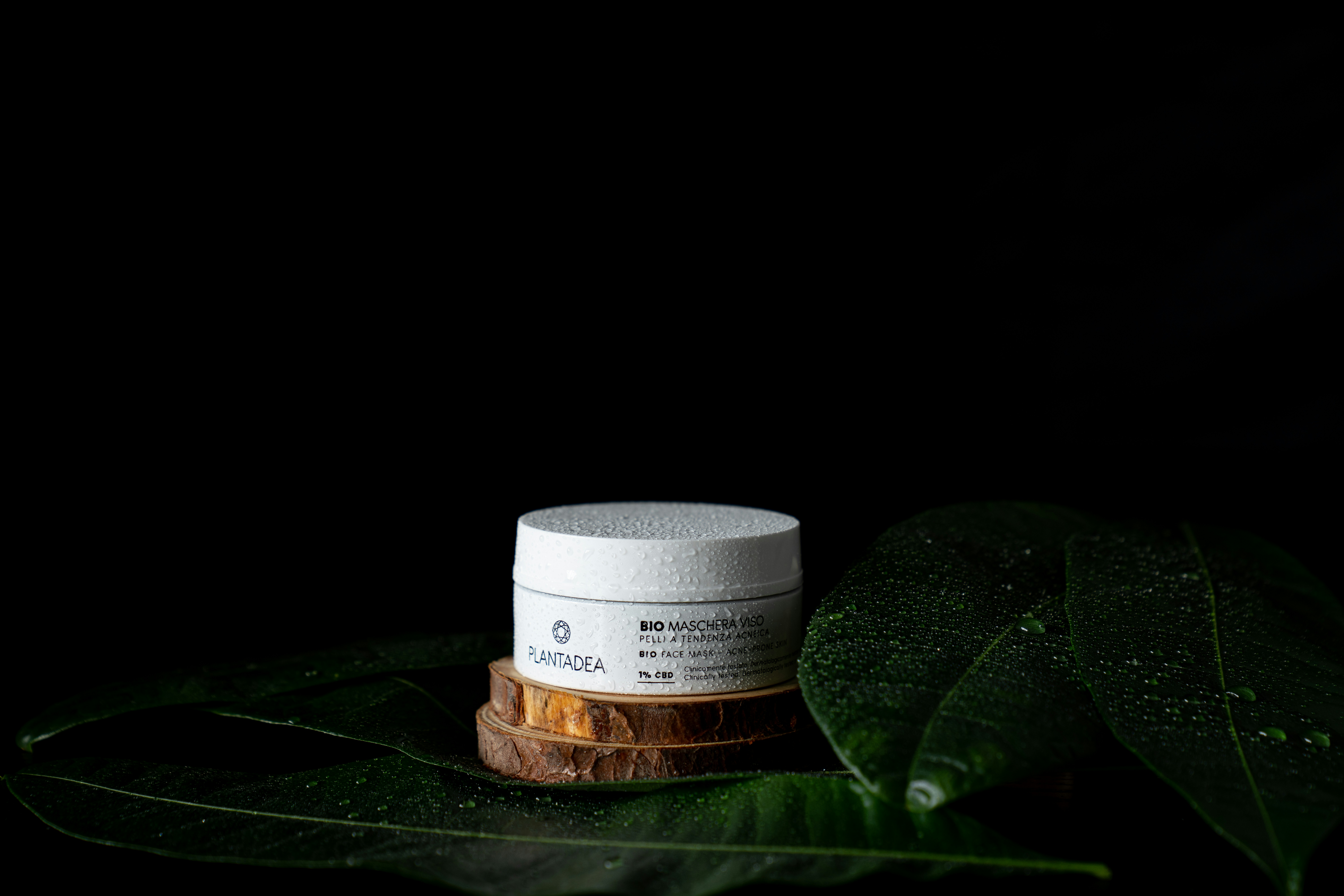 A white cosmetic jar with text sits on a wooden platform surrounded by large green leaves with water droplets on a dark background. The leaves and droplets suggest a natural or organic theme.