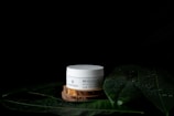 A white cosmetic jar with text sits on a wooden platform surrounded by large green leaves with water droplets on a dark background. The leaves and droplets suggest a natural or organic theme.