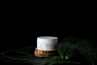 A white cosmetic jar with text sits on a wooden platform surrounded by large green leaves with water droplets on a dark background. The leaves and droplets suggest a natural or organic theme.