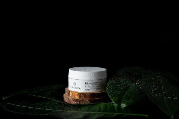 A white cosmetic jar with text sits on a wooden platform surrounded by large green leaves with water droplets on a dark background. The leaves and droplets suggest a natural or organic theme.