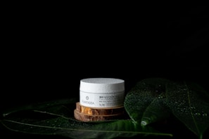 A white cosmetic jar with text sits on a wooden platform surrounded by large green leaves with water droplets on a dark background. The leaves and droplets suggest a natural or organic theme.