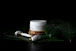 A jar of bio face cream is placed on a wooden coaster surrounded by large, glossy green leaves covered in water droplets. Nearby, a white marble face roller with gold accents rests on the leaves, creating a serene and luxurious spa-like atmosphere.