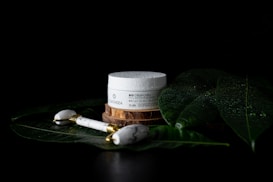 A jar of bio face cream is placed on a wooden coaster surrounded by large, glossy green leaves covered in water droplets. Nearby, a white marble face roller with gold accents rests on the leaves, creating a serene and luxurious spa-like atmosphere.