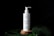 A white pump bottle labeled 'PLANTADEA' is prominently placed on a wooden platform. The bottle has water droplets on its surface, creating a refreshing and natural look. It is surrounded by large green leaves on a black background, enhancing the focus on the product.