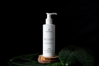 A white pump bottle labeled 'PLANTADEA' is prominently placed on a wooden platform. The bottle has water droplets on its surface, creating a refreshing and natural look. It is surrounded by large green leaves on a black background, enhancing the focus on the product.