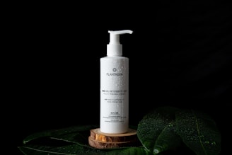 A white bottle of 'Plantadea' bio gel detergent for face cleansing is displayed on a small wooden platform. The bottle has a pump dispenser and is adorned with droplets of water. It is set against a black background with large green leaves, also covered in water droplets, partially visible in the foreground.
