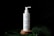 A white bottle of 'Plantadea' bio gel detergent for face cleansing is displayed on a small wooden platform. The bottle has a pump dispenser and is adorned with droplets of water. It is set against a black background with large green leaves, also covered in water droplets, partially visible in the foreground.