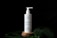 A white bottle of 'Plantadea' bio gel detergent for face cleansing is displayed on a small wooden platform. The bottle has a pump dispenser and is adorned with droplets of water. It is set against a black background with large green leaves, also covered in water droplets, partially visible in the foreground.