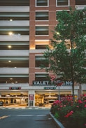A multi-level parking garage with a brick facade and illuminated 'VALET PARKING' sign. There are two signage boards for 'EXIT' and 'ENTER' with red and green lights respectively. A tree with green leaves and a row of red flowering plants are in the foreground. Soft lighting enhances a warm, welcoming atmosphere.