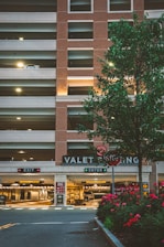 A multi-level parking garage with a brick facade and illuminated 'VALET PARKING' sign. There are two signage boards for 'EXIT' and 'ENTER' with red and green lights respectively. A tree with green leaves and a row of red flowering plants are in the foreground. Soft lighting enhances a warm, welcoming atmosphere.