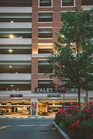 A multi-level parking garage with a brick facade and illuminated 'VALET PARKING' sign. There are two signage boards for 'EXIT' and 'ENTER' with red and green lights respectively. A tree with green leaves and a row of red flowering plants are in the foreground. Soft lighting enhances a warm, welcoming atmosphere.