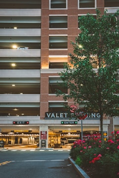 A multi-level parking garage with a brick facade and illuminated 'VALET PARKING' sign. There are two signage boards for 'EXIT' and 'ENTER' with red and green lights respectively. A tree with green leaves and a row of red flowering plants are in the foreground. Soft lighting enhances a warm, welcoming atmosphere.