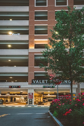A multi-level parking garage with a brick facade and illuminated 'VALET PARKING' sign. There are two signage boards for 'EXIT' and 'ENTER' with red and green lights respectively. A tree with green leaves and a row of red flowering plants are in the foreground. Soft lighting enhances a warm, welcoming atmosphere.