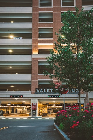 A multi-level parking garage with a brick facade and illuminated 'VALET PARKING' sign. There are two signage boards for 'EXIT' and 'ENTER' with red and green lights respectively. A tree with green leaves and a row of red flowering plants are in the foreground. Soft lighting enhances a warm, welcoming atmosphere.