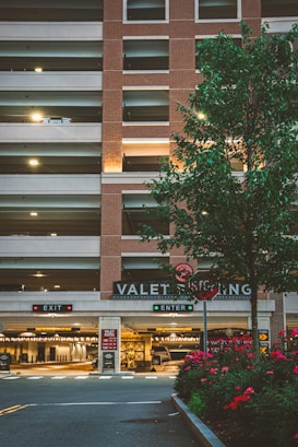 A multi-level parking garage with a brick facade and illuminated 'VALET PARKING' sign. There are two signage boards for 'EXIT' and 'ENTER' with red and green lights respectively. A tree with green leaves and a row of red flowering plants are in the foreground. Soft lighting enhances a warm, welcoming atmosphere.