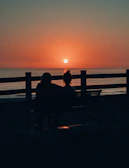 A couple seated on a charcoal-colored bench, sharing a quiet conversation as the sun sets behind them.