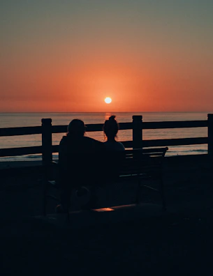 A couple seated on a charcoal-colored bench, sharing a quiet conversation as the sun sets behind them.