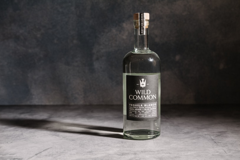 A bottle of Wild Common Tequila Blanco stands upright on a surface against a dark, textured background. The label indicates it is made from 100% agave, specifically Blue Weber agave.
