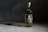 A bottle of Wild Common Tequila Reposado is placed on a smooth, dark surface with subtle lighting creating a shadow. The bottle has a green label with white text, displaying details about the tequila including '100% Agave' and 'Blue Weber.' The background is a textured, dark gray to black backdrop, enhancing the focus on the bottle.