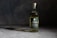A bottle of Wild Common Tequila Reposado is placed on a smooth, dark surface with subtle lighting creating a shadow. The bottle has a green label with white text, displaying details about the tequila including '100% Agave' and 'Blue Weber.' The background is a textured, dark gray to black backdrop, enhancing the focus on the bottle.