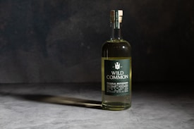 A bottle of Wild Common Tequila Reposado is placed on a smooth, dark surface with subtle lighting creating a shadow. The bottle has a green label with white text, displaying details about the tequila including '100% Agave' and 'Blue Weber.' The background is a textured, dark gray to black backdrop, enhancing the focus on the bottle.