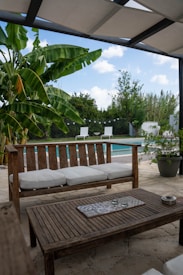 A cozy outdoor patio area features a wooden bench and coffee table with white cushions, surrounded by lush greenery. In the background, there is a swimming pool with two white lounge chairs, enclosed by a fence. The overhead covering provides shade, and a few decorative candles are placed on the table.