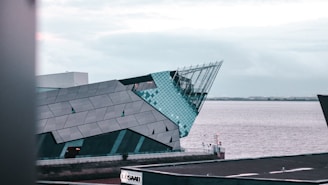 A modern architectural building with sharp angles, featuring a combination of metal and glass elements. The structure is situated near a body of water, with a cloudy sky overhead. In the foreground, there is a visible advertisement sign for Saab.